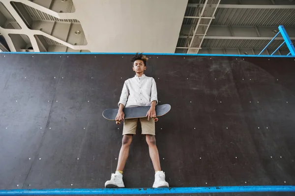 スケートボードを保持している若い黒人のティーンエイジャーの広い角度ショットと都市のスケートパークでランプに立っている間カメラを見て コピースペース — ストック写真
