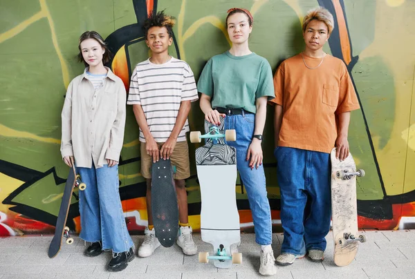 グラフィティ ウォールに立ち向かうスケートボードと 都市のストリート セッティングでカメラを見るティーンエイジャーの多様なグループの活発なショット — ストック写真