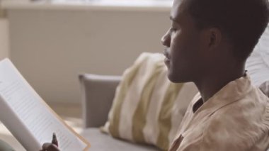 Side view chest up slowmo of young African American man smoking electronic cigarette while reading business documents in office