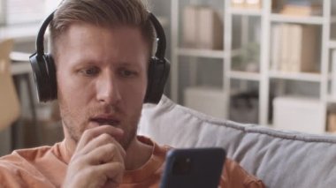 Medium closeup portrait of young Caucasian man listening to music in headphones, scrolling on smartphone and smoking electronic cigarette, sitting on sofa at home