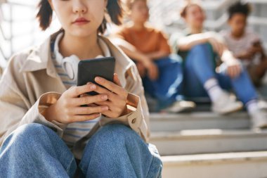 Metal merdivenlerde arka planda bir grup arkadaşla otururken akıllı telefon tutan genç kızın yakın plan fotoğrafı.