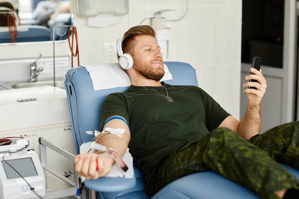 Portrait of smiling military man giving blood while laying in chair at blood donation center and holding smartphone, copy space