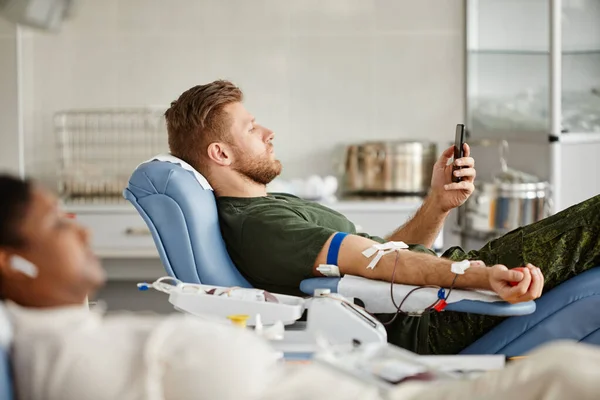 军人躺在献血中心的椅子上和智能手机上看视频时献血的侧视图 — 图库照片