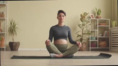 Slowmo portrait of young pregnant woman in tight activewear sitting in lotus pose on yoga mat at home meditating