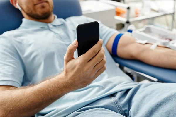 在医学中心舒适的椅子上躺着 拿着智能手机献血的年轻人的近照 — 图库照片