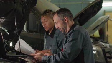 Two diverse male and female mechanics looking at blueprint while inspecting camper van standing under its hood in car service