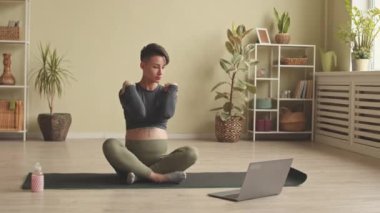Slowmo of contemporary young pregnant woman having online sports training from home via laptop, sitting on yoga mat and repeating after coach