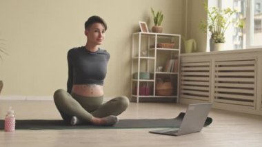 Slowmo of young slim pregnant woman doing physical and stretching exercises on yoga mat at home while watching online training on laptop