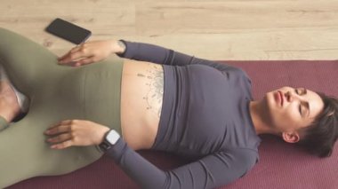 Top view of relaxed pregnant woman lying on yoga mat with her arms stretched, having light yoga practice at home