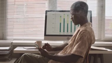 Young Black man sitting in front of window at office desk using smartphone and vaping