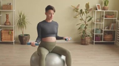 Energetic young pregnant woman exercising with dumbbells while sitting on fit ball having indoor fitness training