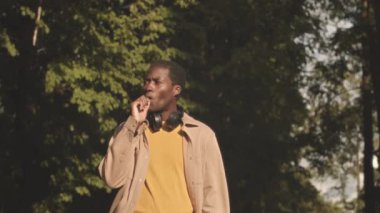 Medium slowmo of relaxed young Black man smoking e cigarette while walking slowly in park on sunny day