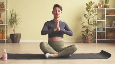 Slowmo of modern young pregnant woman with short haircut and tattoo on her belly meditating on yoga mat at home