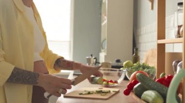 Cropped shot of tattooed pregnant woman in oversized light ye3llow shirt making healthy vegetable salad at bright modern kitchen
