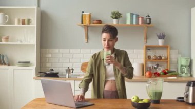 Medium shot of contemporary pregnant woman with short haircut and tattoos using laptop and drinking vitamin green smoothie at kitchen