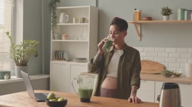 Medium portrait of healthy young pregnant woman with short haircut and tattoos drinking homemade green smoothie at kitchen and smiling at camera