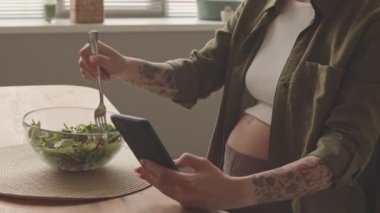 Cropped shot of tattooed woman expecting baby texting on smartphone while eating green salad having lunch at home