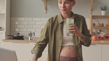 Tilt shot of modern pregnant woman with short dark hair and tattoos on her arms and belly working on laptop from home drinking vitamin green smoothie