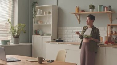 Medium shot of young pregnant woman with short haircut and tattoos standing at kitchen eating vitamin green salad