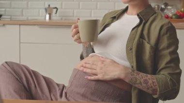 Cropped slowmo of tattooed pregnant woman stroking her belly tenderly while sitting at wooden kitchen table