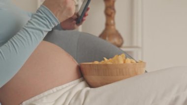 Close up of unrecognizable pregnant woman eating chips while scrolling on smartphone indoors