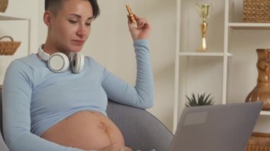 Young Caucasian pregnant woman eating sweet biscuit waffles while working on laptop from home