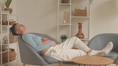Short haired young pregnant woman with hands on her belly relaxing in armchair at home