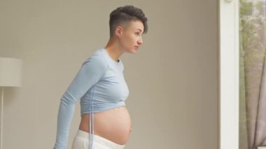 Medium shot of short haired young pregnant woman measuring her tummy while standing by window at home