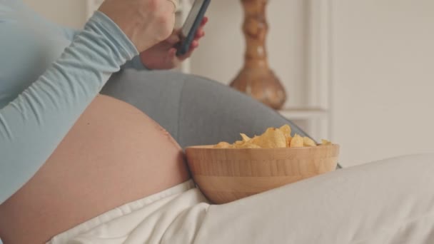 Close Unrecognizable Pregnant Woman Eating Chips While Scrolling Smartphone Indoors — стокове відео