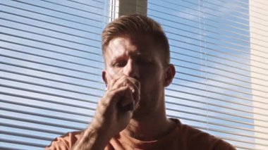 Medium closeup portrait of young Caucasian man vaping, standing against clear blue sky from window