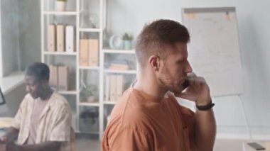 Chest up slowmo of young Caucasian man smoking electronic cigarette while talking on smartphone in office