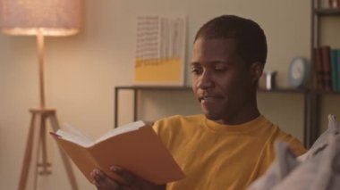 Chest up of young Black man smoking electronic cigarette while reading exciting book at home in evening