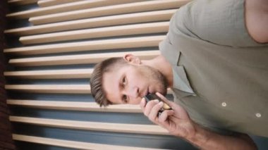 Vertical pov shot of young Caucasian man vaping and talking at camera outdoors