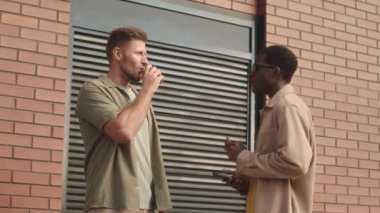 Medium slowmo of two diverse male friends talking and smoking electronic devices outdoors in urban environment