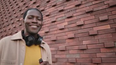 Chest up portrait of smiling young Black hipster man with electronic cigarette looking at camera standing against brick wall outdoors