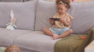 Güzel Afrikalı Amerikalı kız akıllı telefondan video izliyor oturma odasındaki gri koltukta oturuyor babası da yakında oturan bebekle oynuyor.
