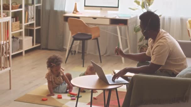 带着耳机的非裔美国人在家里的笔记本电脑上工作 而他的两个可爱的幼儿则在附近玩玩具 — 图库视频影像