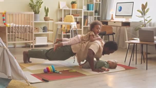 活泼的非洲裔美国男子 两个蹒跚学步的孩子的父亲在家里做俯卧撑 女儿躺在他的背上 儿子在他的怀里 — 图库视频影像