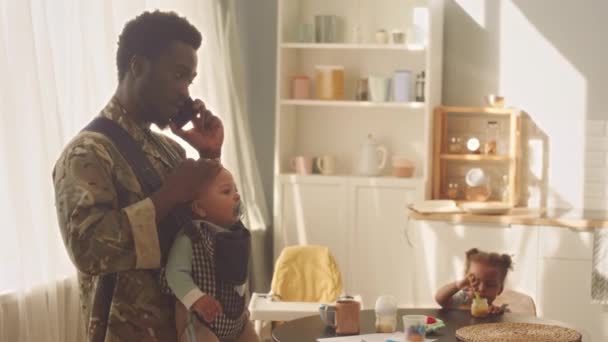 バックパックキャリアの赤ん坊の息子と黒人の男は キッチンで携帯電話で話しながら 彼の幼い娘は背景でテーブルでピューレを食べています — ストック動画