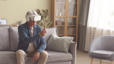 VR kulaklıklı beyaz adam oturma odasında kanepede oturuyor, video oyunları oynuyor, video kumandasındaki düğmeye basıyor ve kafasını farklı yönlere çeviriyor.