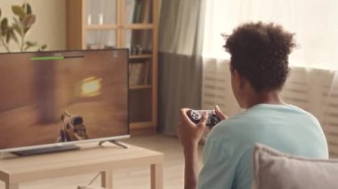 Oturma odasında FPS video oyunu oynarken kontrol cihazı kullanan genç bir çocuğun arka görünümü