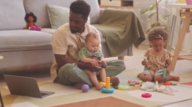 Bekar Afrikalı Amerikalı iki çocuk babası. Oturma odasında halı üzerinde oyuncaklarla oynarken evden çalışmaya çalışıyor.