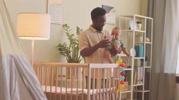 自宅で彼のかわいい赤ちゃんの息子をロックしながらスマートフォンを使用して幸せな若いアフリカ系アメリカ人男性 — ストック動画