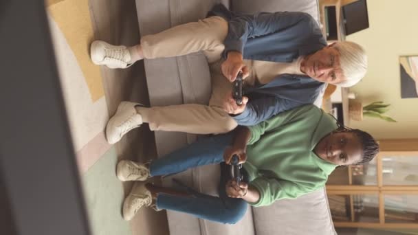 リビングルームでソファーでビデオゲームをしながら楽しんでいる2人の多世代の友人の垂直ショット — ストック動画