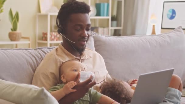 年轻的非洲裔美国人头戴耳机 带着话筒 在家里的笔记本电脑上工作 照顾着身边躺在沙发上的两个幼儿 — 图库视频影像