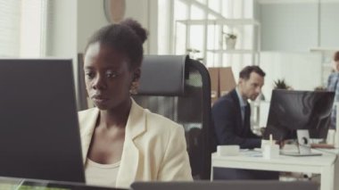 Genç Afrikalı Amerikalı kadın programcının orta yavaş portresi. Kameraya gülümsüyor. Çalışma masasında bilgisayar monitörünün önünde oturuyor.
