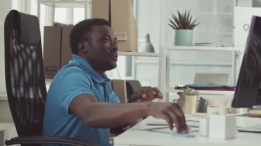 Mavi polo tişörtlü genç siyahi adam modern bilişim ofisinde bilgisayarla çalışıyor.
