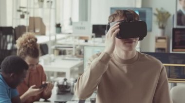 VR kulaklıklı genç beyaz adam modern bilişim ofisinde çeşitli meslektaşlarıyla çalışırken elleriyle hareketler yapıyor.