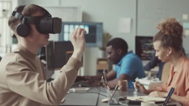 Genç erkek yazılım mühendisi VR ile modern ofiste çalışırken çeşitli meslektaşları bilgisayarlarında kodlama yapıyor.