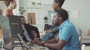 Genç Siyahi Adam 'ın arka planda çok ırklı iş arkadaşlarıyla sohbet ederken bilgisayar programında kod yazmasını yavaşlat, başarılı bilişim şirketinde hep birlikte çalışsınlar.